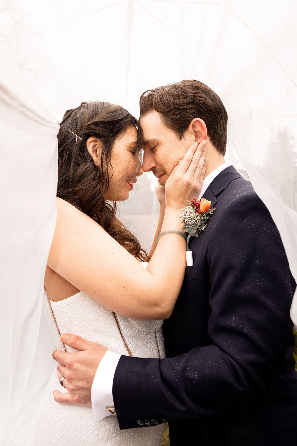 Bride and groom share a tender forehead touch, embracing under a white veil on their wedding day.