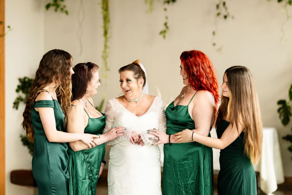 Bride in white lace gown surrounded by four bridesmaids in emerald green satin dresses during wedding preparations.