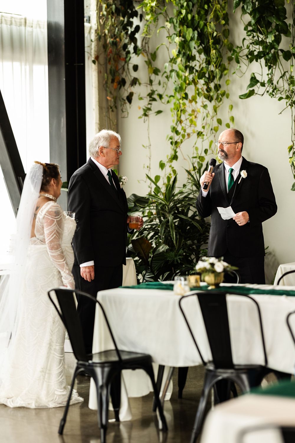 Officiant reads vows at elegant indoor wedding ceremony with bride and groom surrounded by lush greenery.