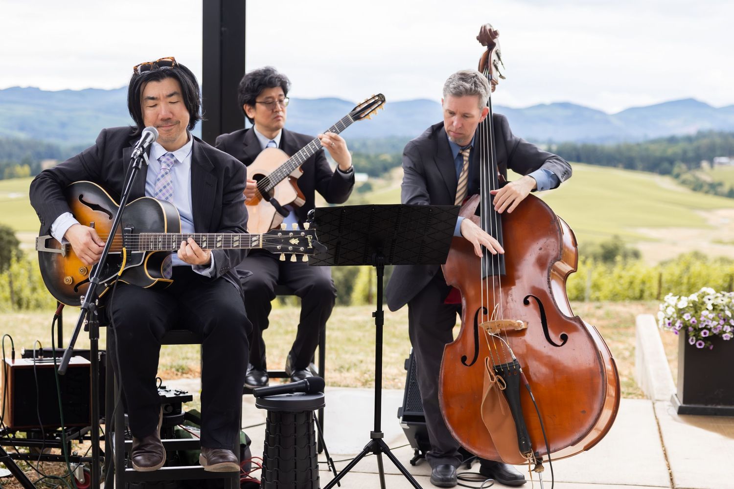 Musicians perform with acoustic instruments including guitar and upright bass at an outdoor venue with mountain views.