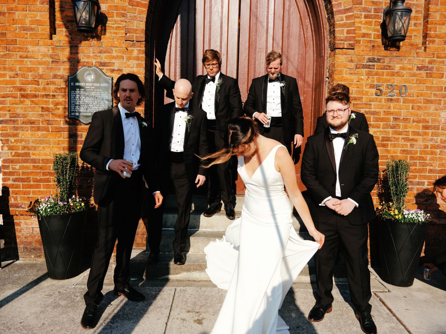 Wedding party in formal attire poses playfully while walking along a brick wall exterior.