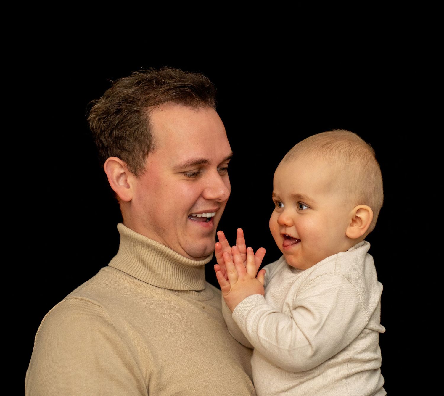 Een lachende vader houdt zijn speelse baby vast, die in zijn handjes klapt, tegen een zwarte achtergrond.