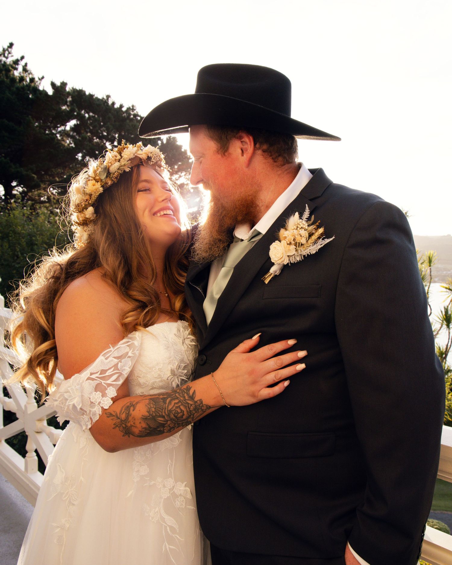 A bride in a floral crown and groom in a black cowboy hat share a joyful moment on their wedding day.
