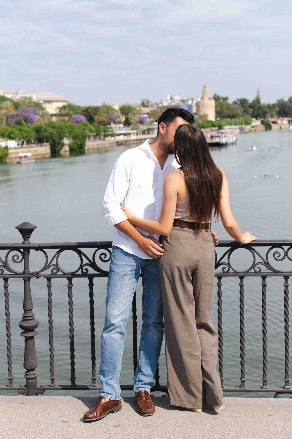 Romantic couple sharing a kiss on a scenic riverside bridge with lush greenery and historic tower in background.