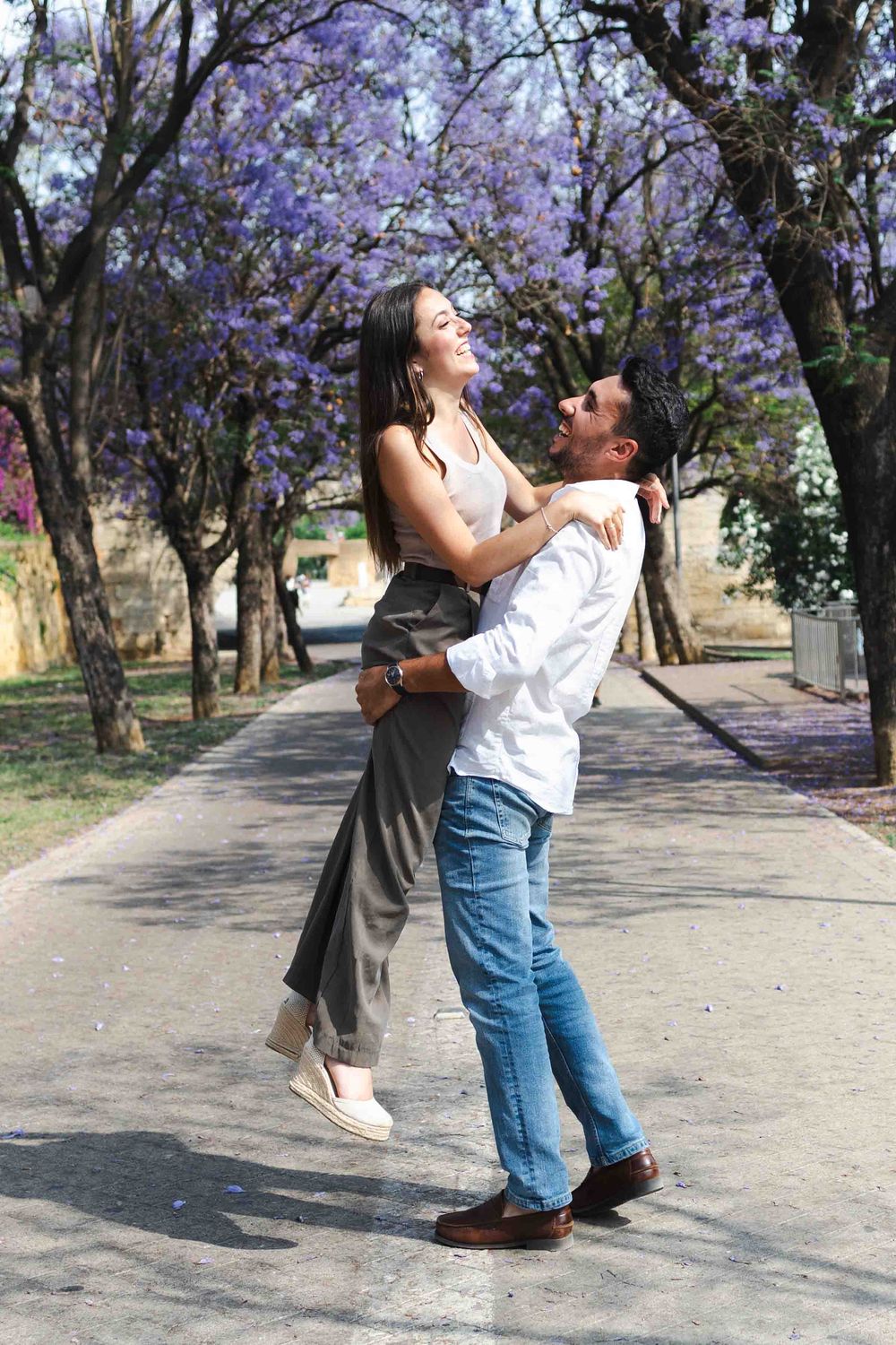 Couple embracing under blooming jacaranda trees on a sunny path, man lifting woman as they share a joyful moment.