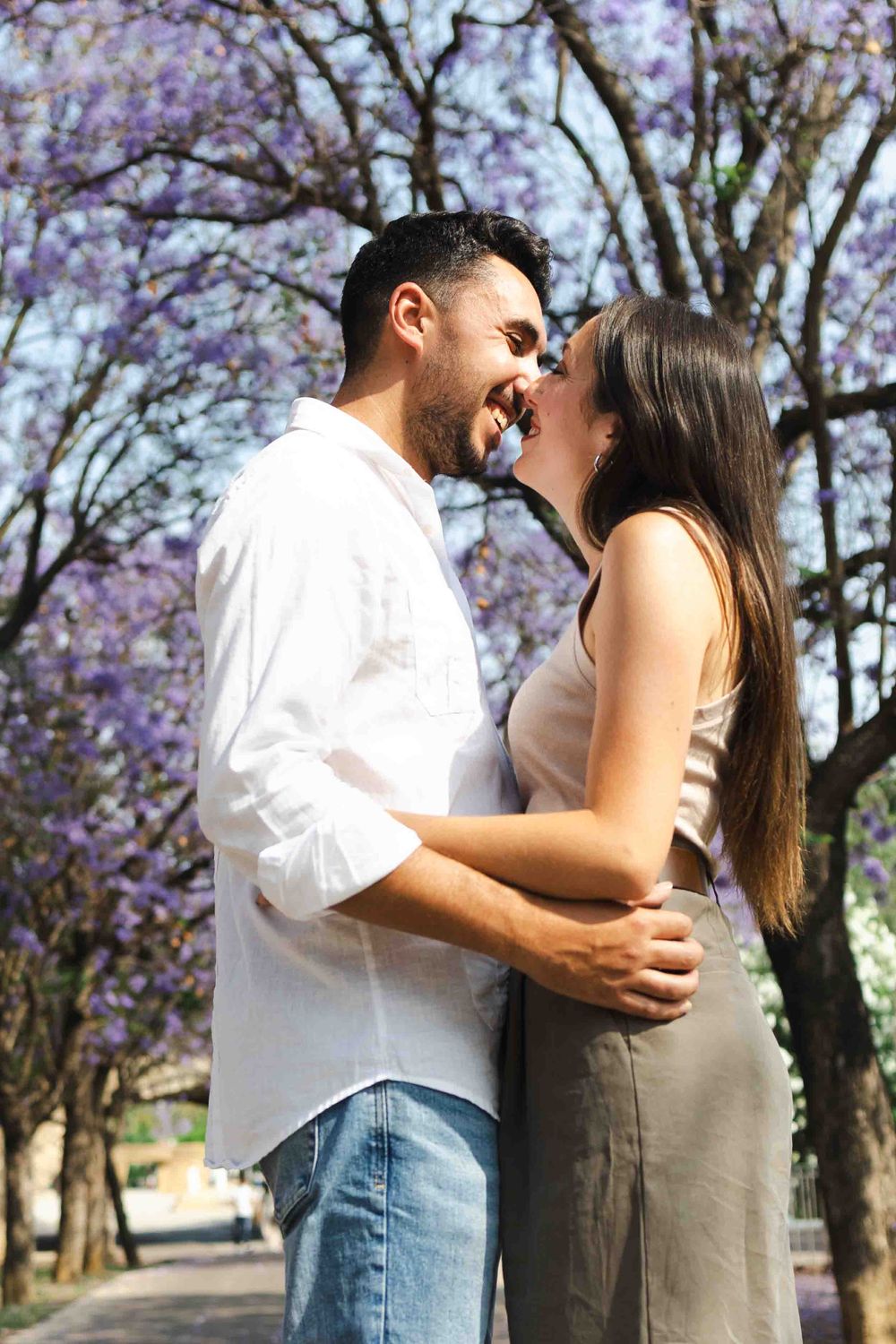 Romantic couple embracing and almost kissing outdoors under blooming purple jacaranda trees on a sunny day.
