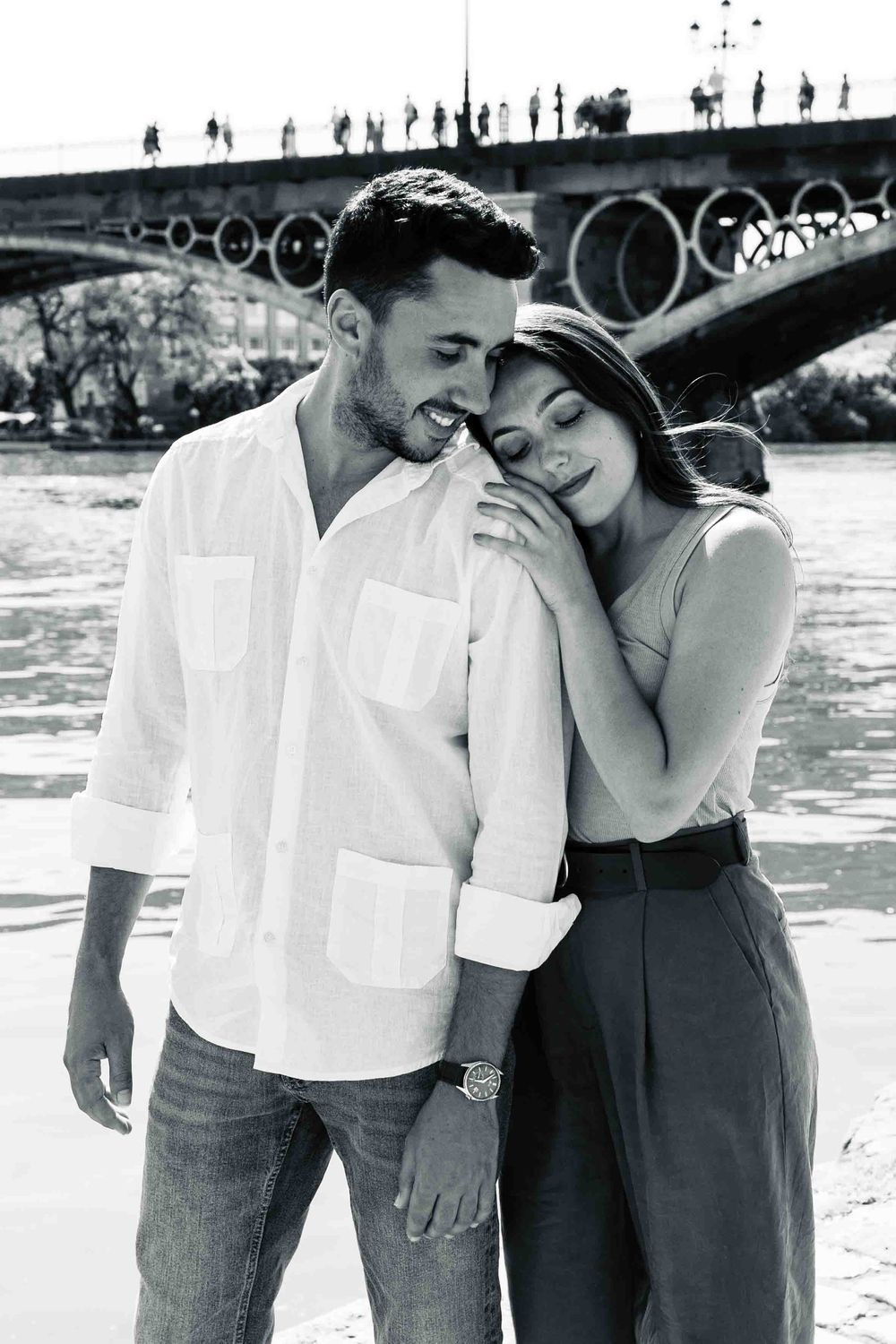 Black and white photo of a romantic couple embracing near a river with a bridge in the background.