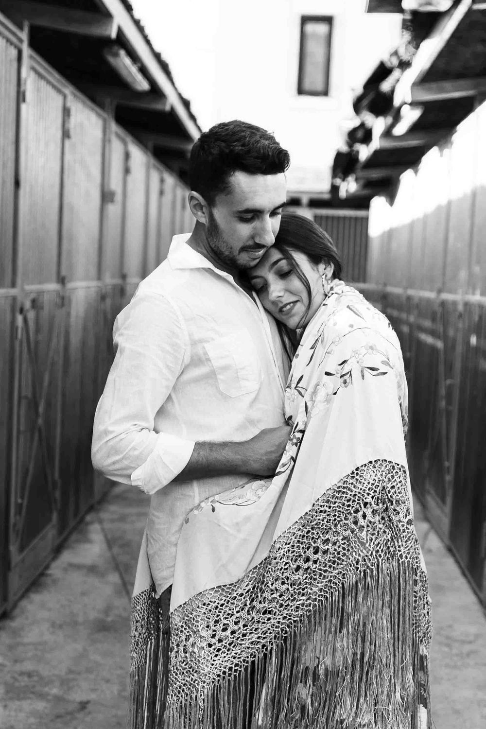 Black and white photo of a romantic couple embracing tenderly, woman wrapped in a fringed shawl, leaning on mans chest.