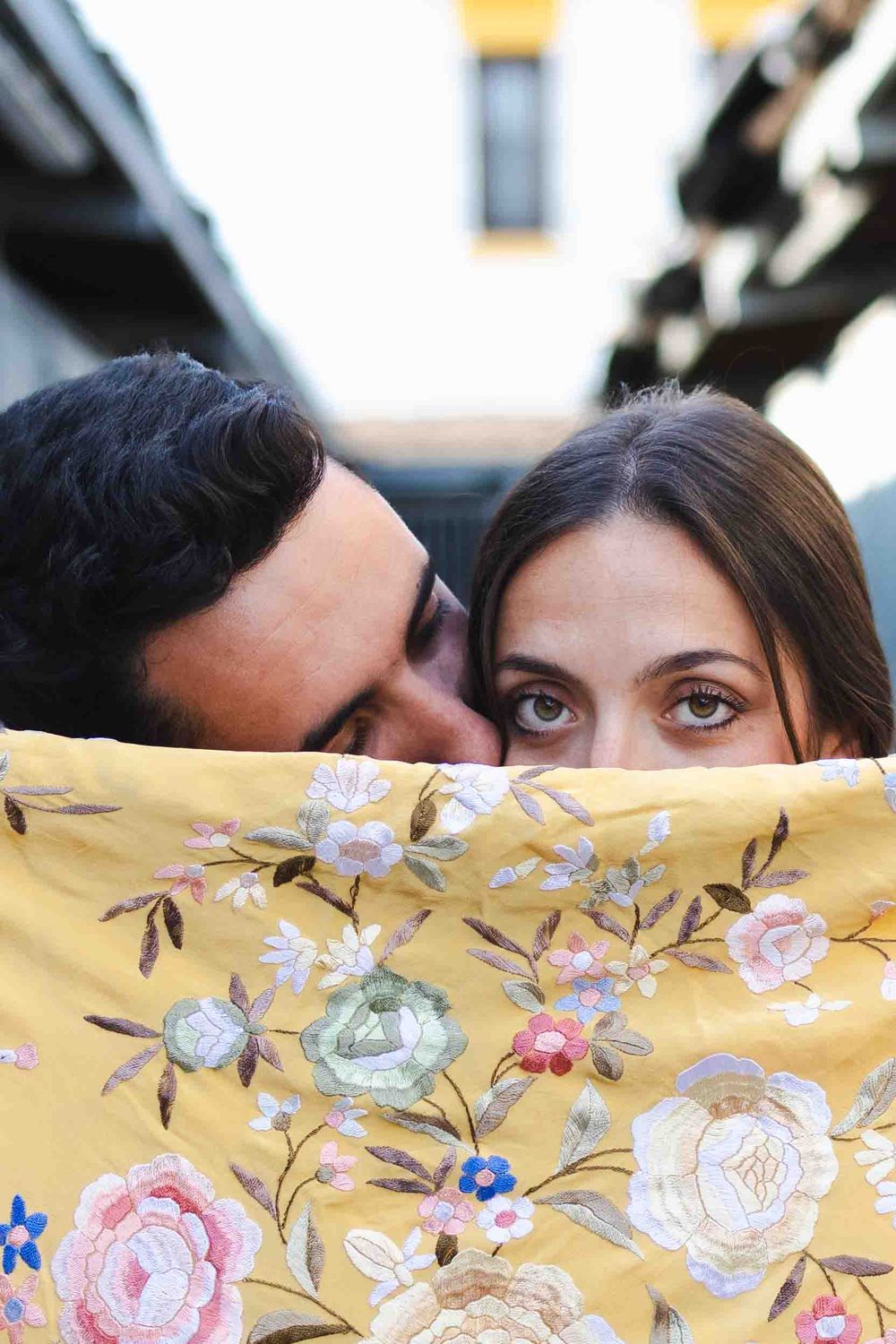 A couple playfully hides behind a yellow floral fabric, showing only their eyes and foreheads.