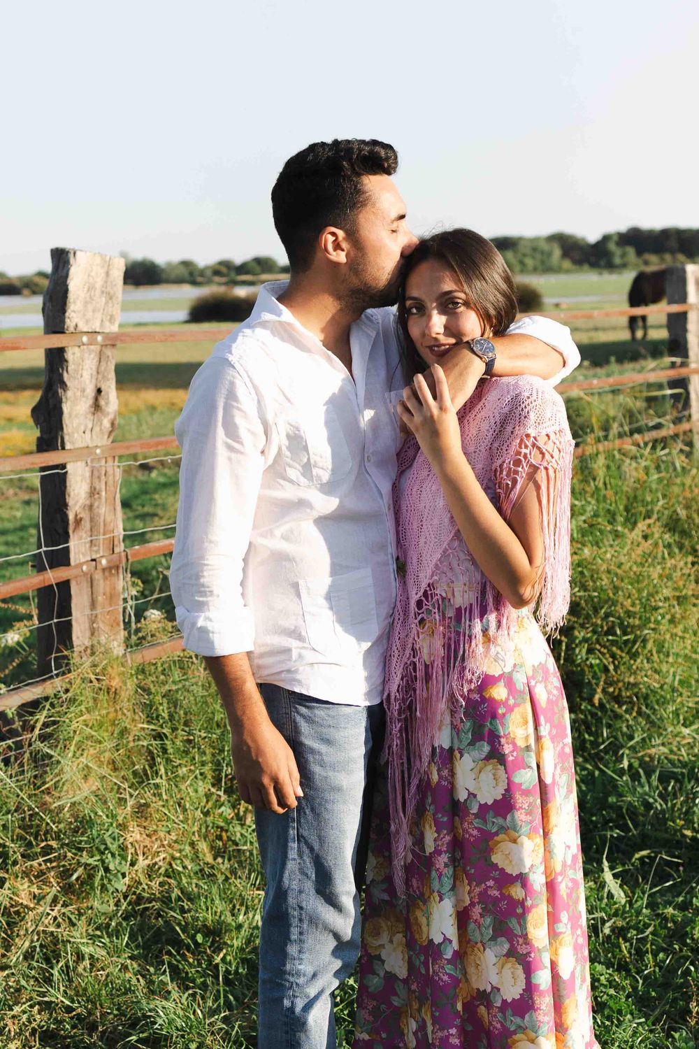Romantic couple embracing outdoors near a rustic wooden fence in a green field at golden hour.