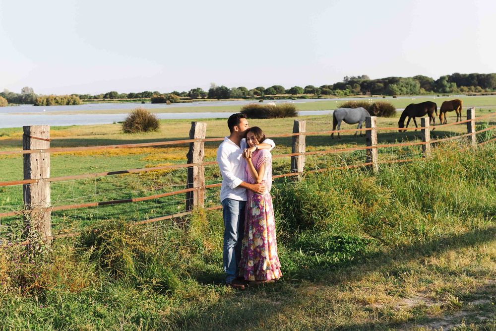 Couple embracing by a wooden fence in a green pasture with horses grazing near a lake at golden hour.
