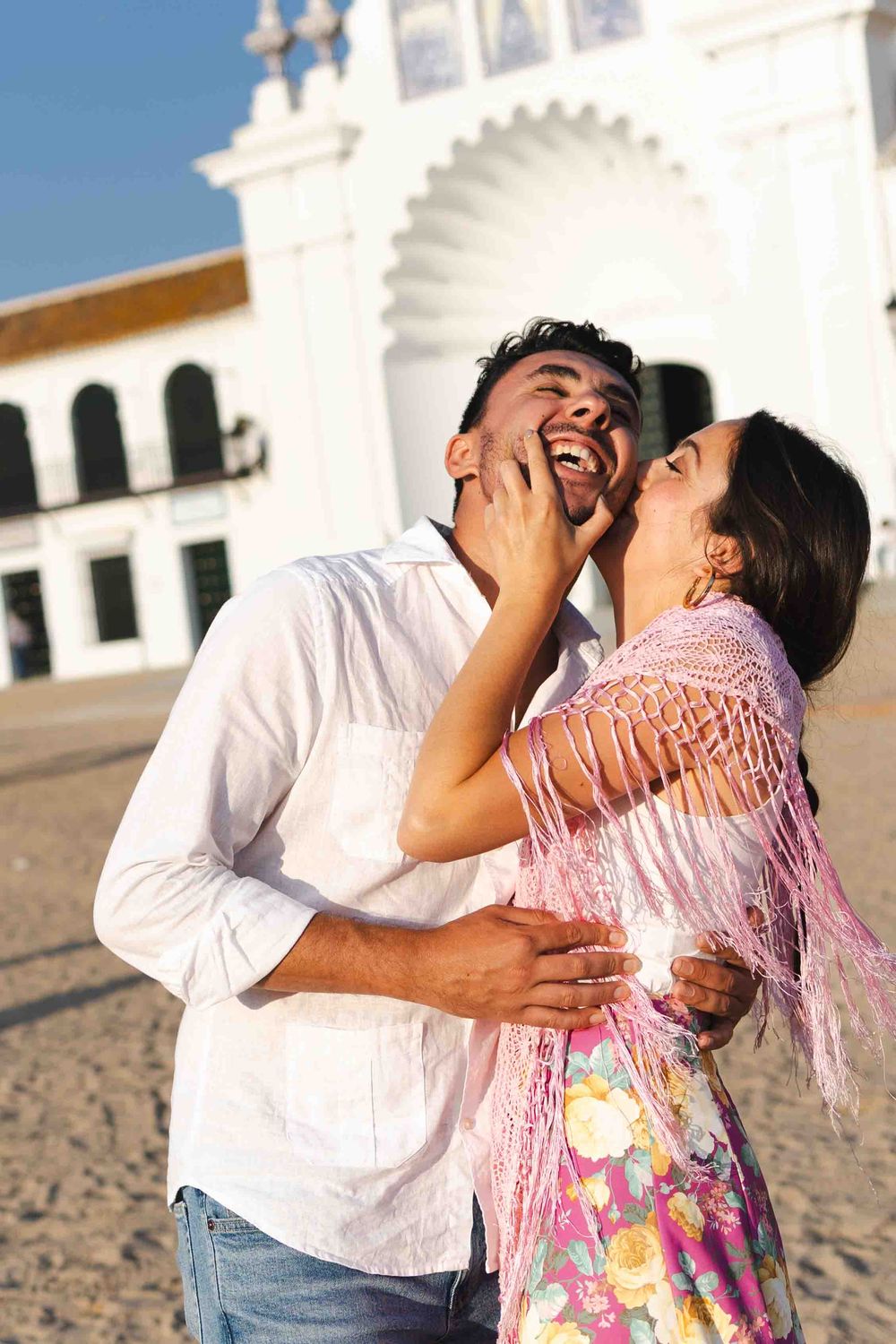 A joyful couple laughing and embracing outdoors near a white colonial building on a sunny day.