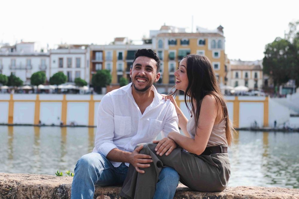 Happy couple sitting together by a riverside in Seville, Spain, laughing and enjoying a sunny day outdoors.