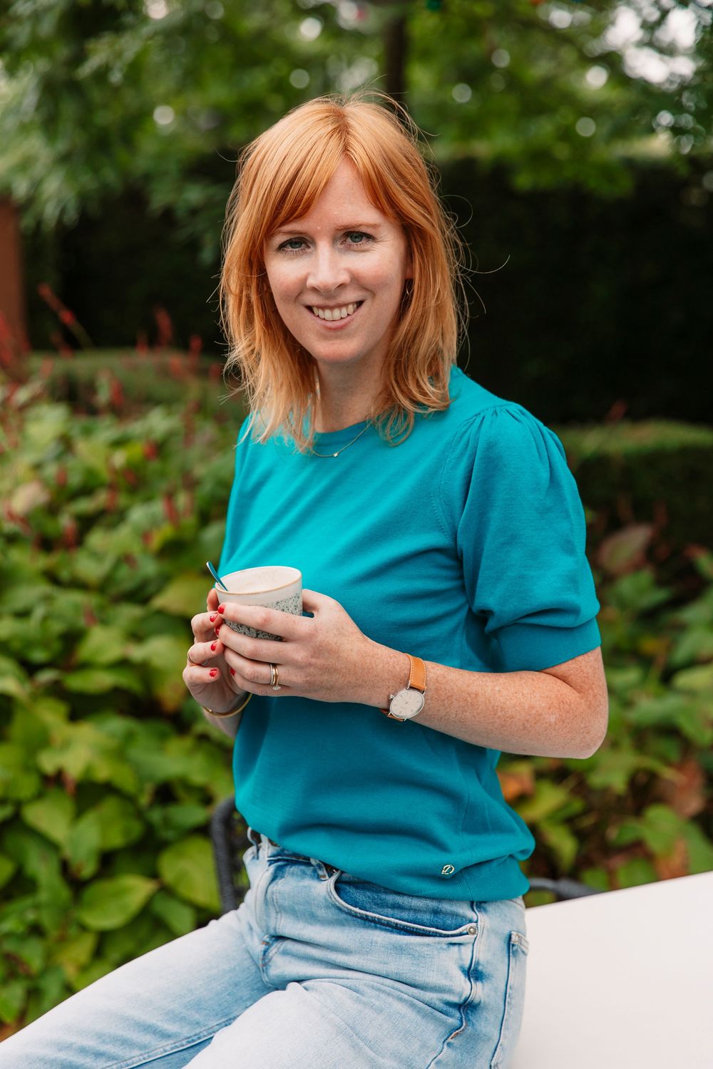 Person in teal sweater holding coffee cup against garden backdrop.