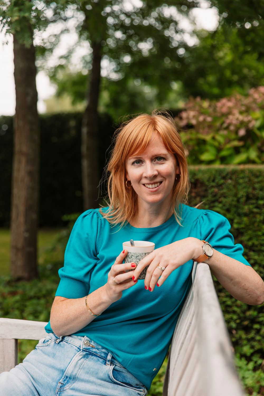 Person in teal top and jeans relaxing on white outdoor bench with beverage.