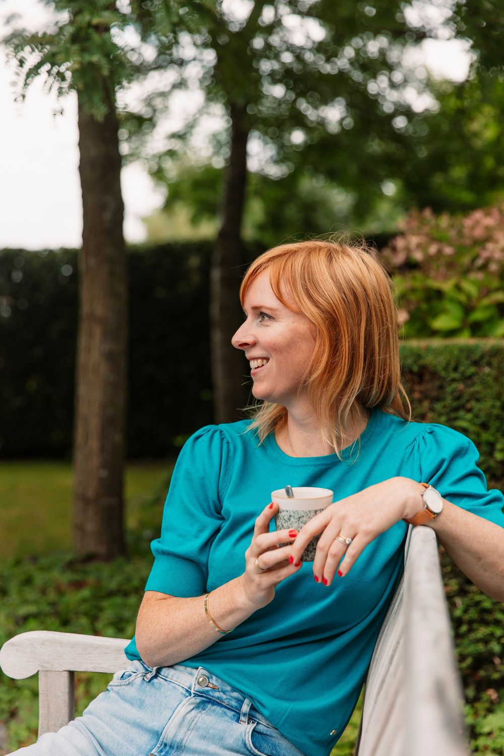 Person in teal shirt enjoying coffee outdoors on white garden bench.