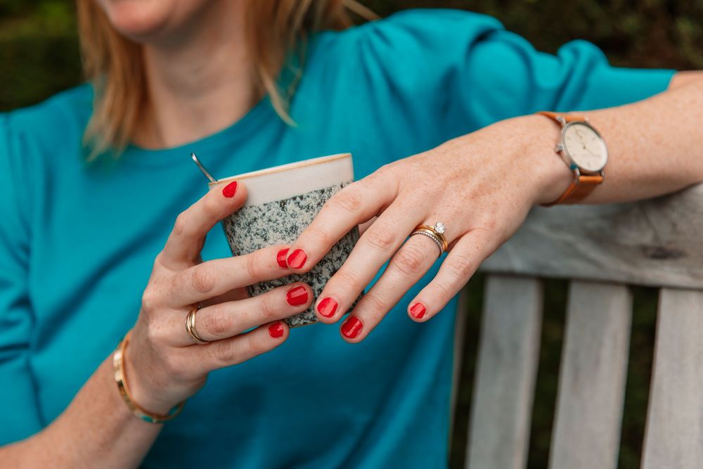 Close-up of hands with red nail polish holding ceramic cup, wearing teal sweater.