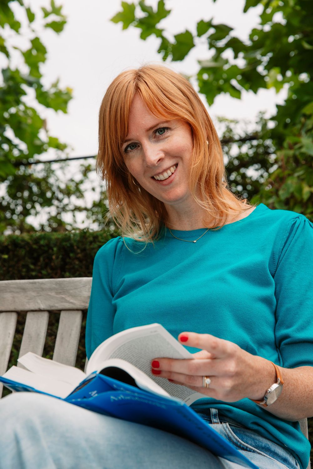 Person in turquoise shirt reading book while relaxing on garden bench.