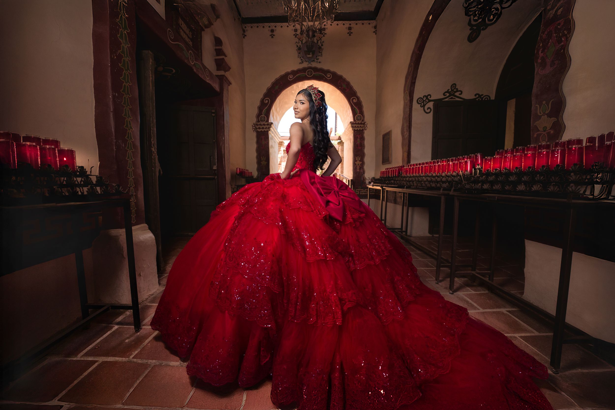 A person in a flowing red ball gown poses dramatically in a dimly lit hallway with arched architecture.