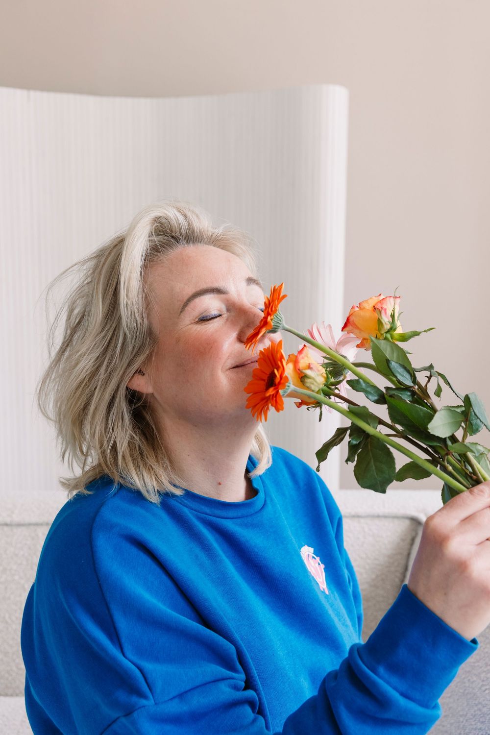Person in bright blue sweater smelling orange and pink flowers.