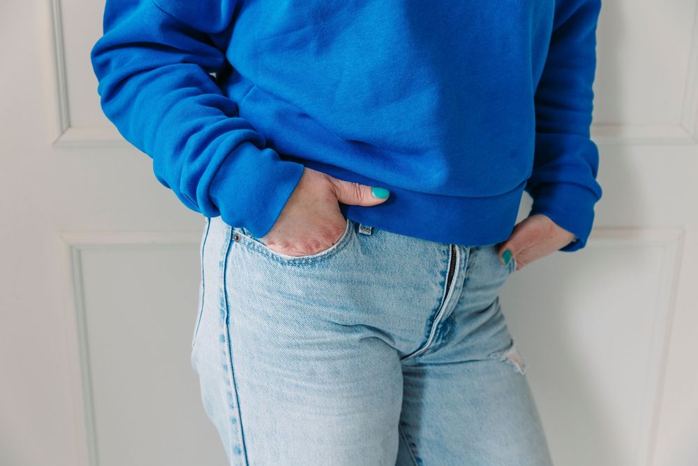 Person wearing light blue jeans and a bright blue sweater standing against a white wall.