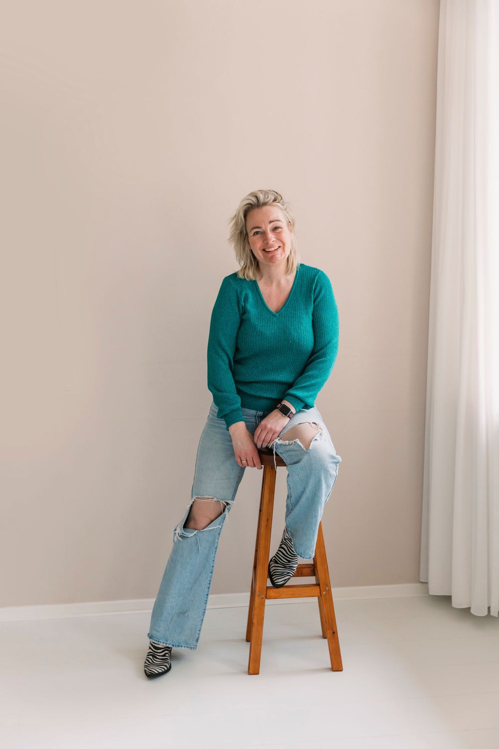 Casual poses on wooden stool wearing teal sweater and light denim.