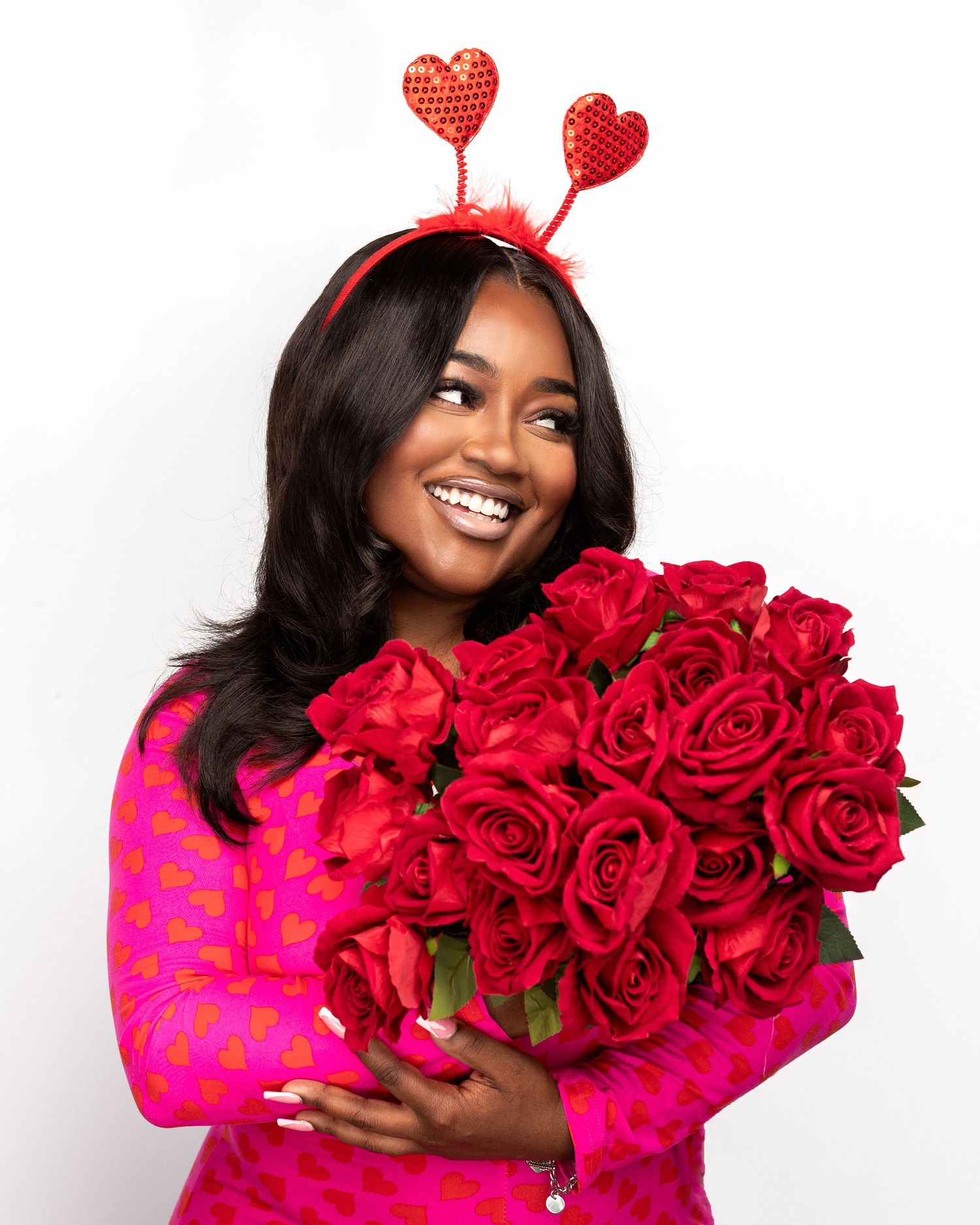 Someone wearing a bright pink sweater holds a large bouquet of vibrant red roses against a white background.