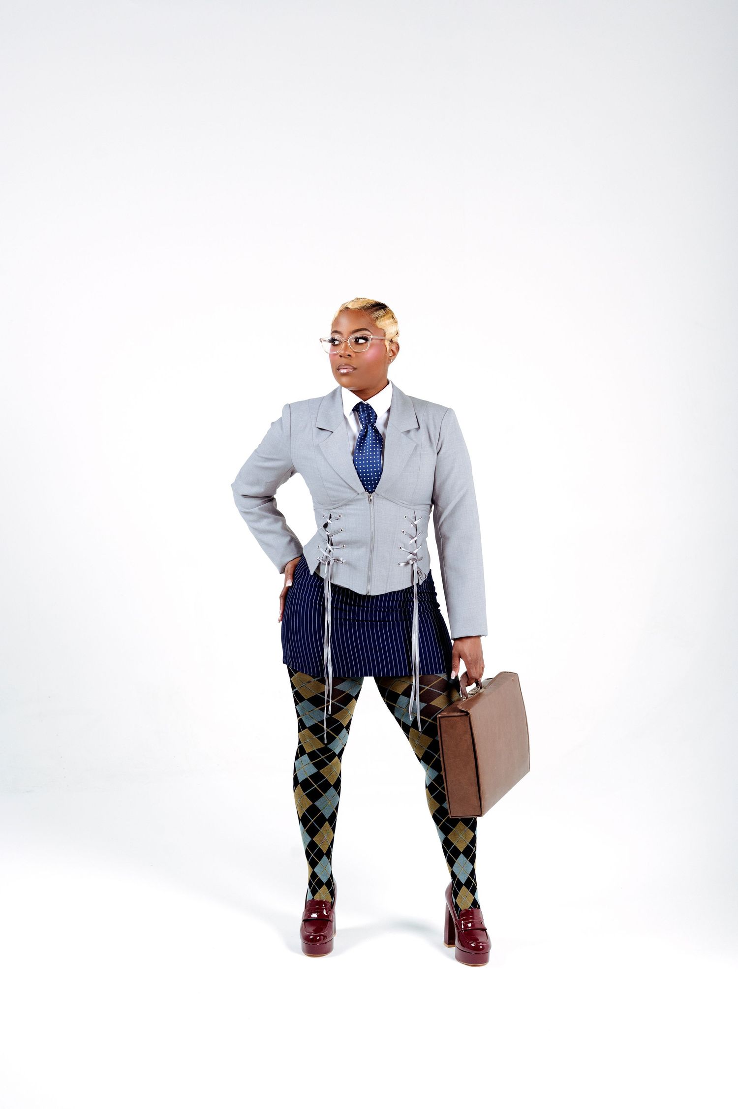 Person in gray blazer, blue tie, argyle tights, and burgundy shoes holding a briefcase against white background.