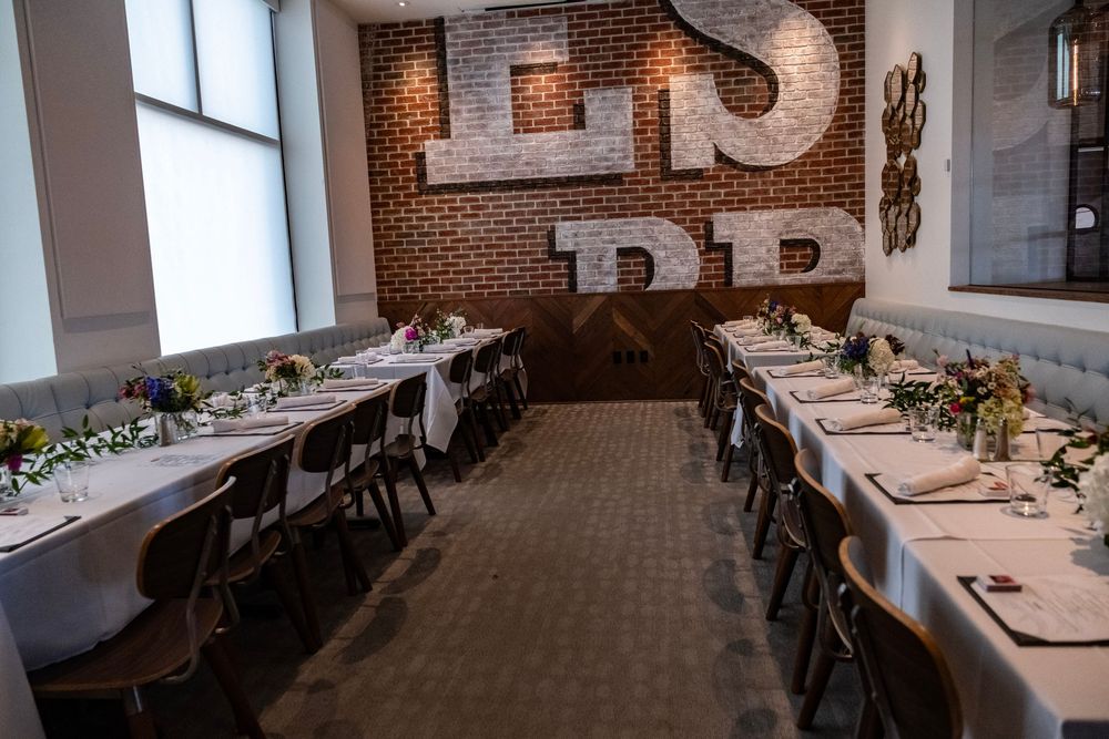 An elegant private dining room with long white-clothed tables, brick walls, and modern decor for a special event setup.