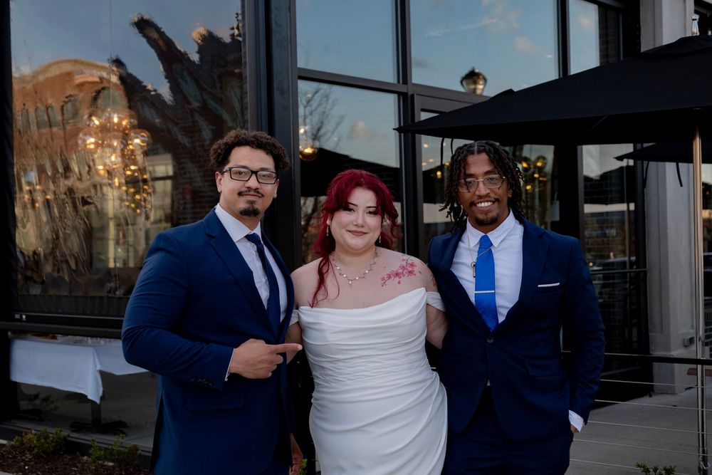 Three wedding guests in formal attire pose together outside a venue with glass windows at dusk.
