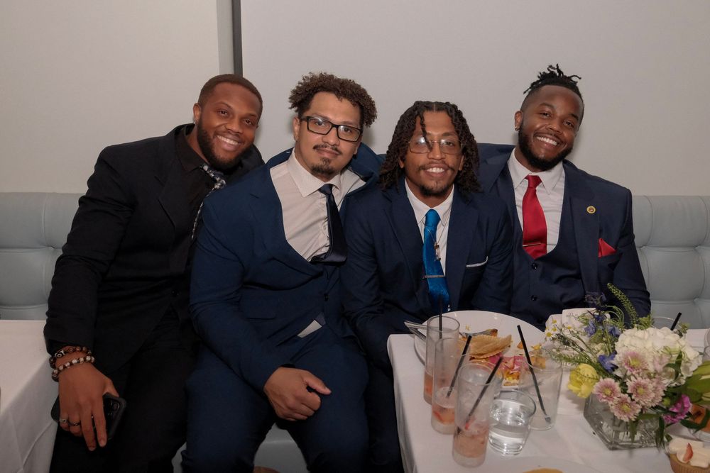 Four people in formal suits sit together at a table with floral centerpiece decorations at an event.