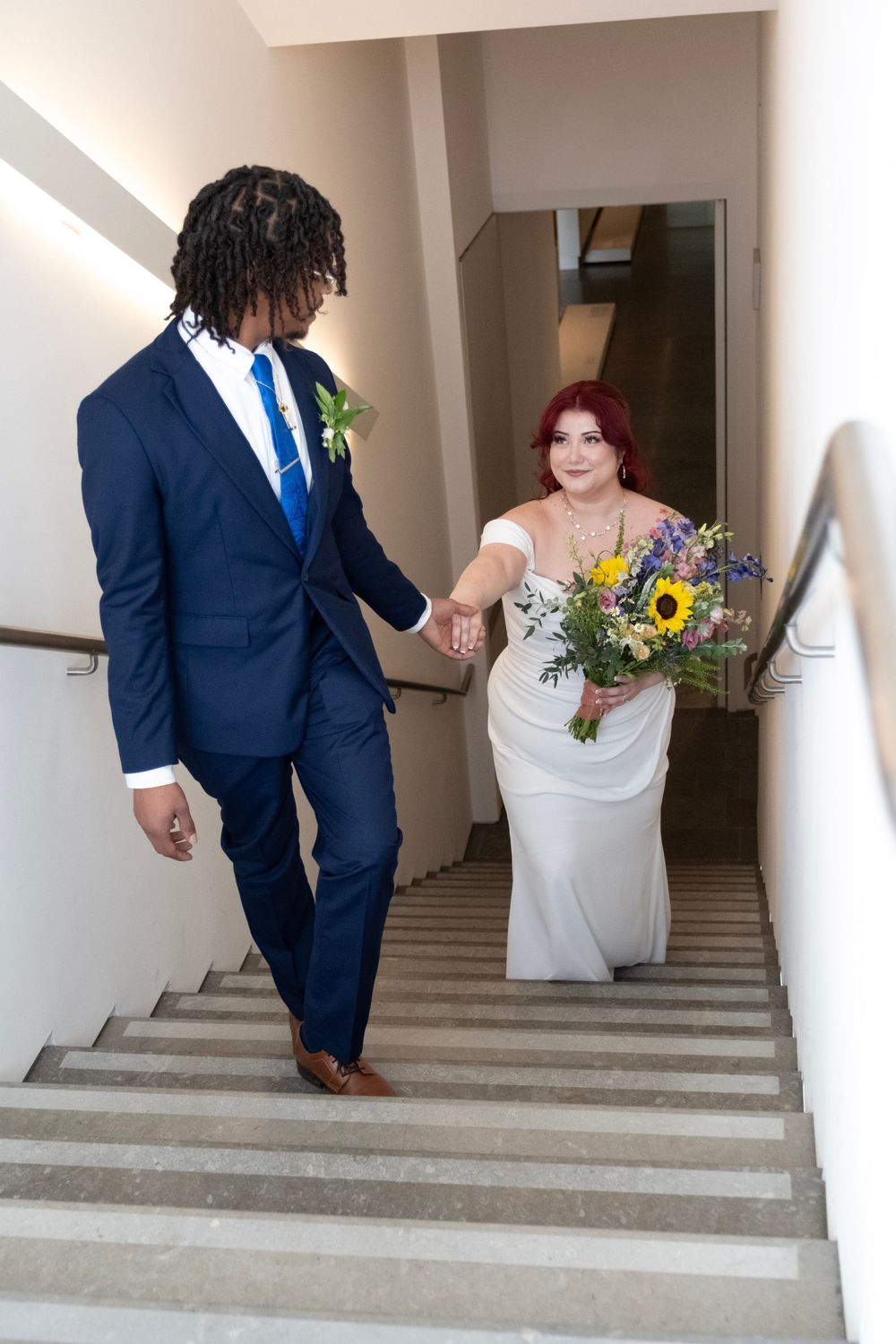 Wedding couple descends white stone stairs in formal attire with sunflowers and blue accents.