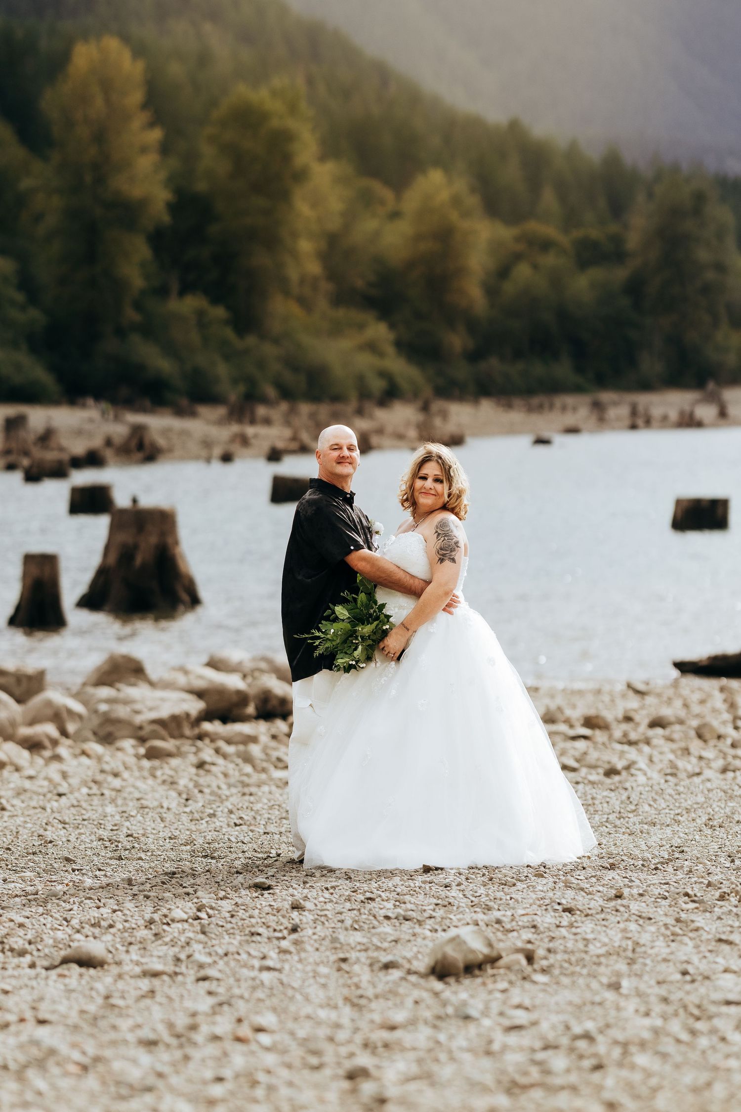 A couple in wedding attire sits together on a rocky beach shore with tree-lined mountains in the background.