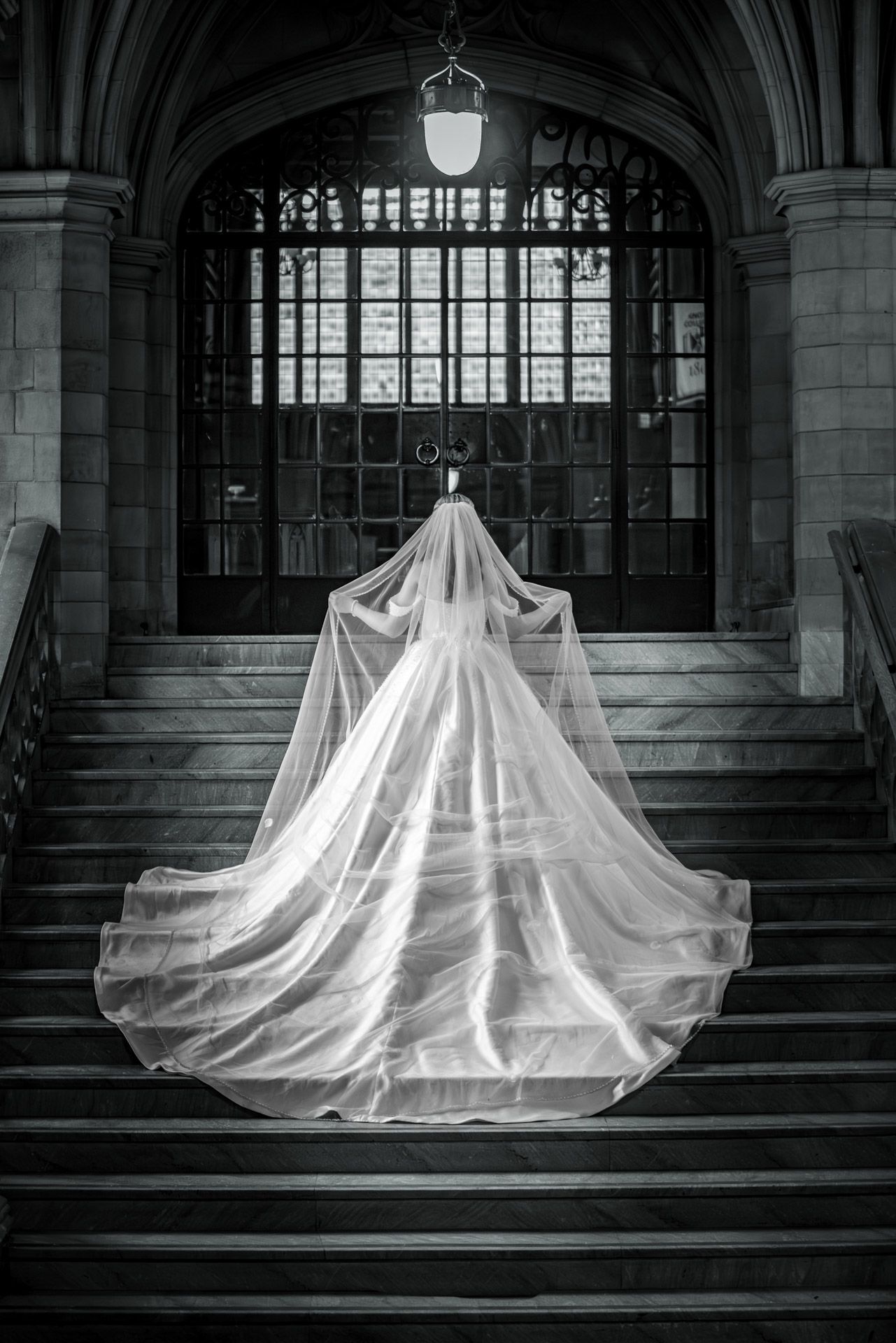 Bride in flowing white gown and veil ascending grand stone staircase in black and white wedding photo.