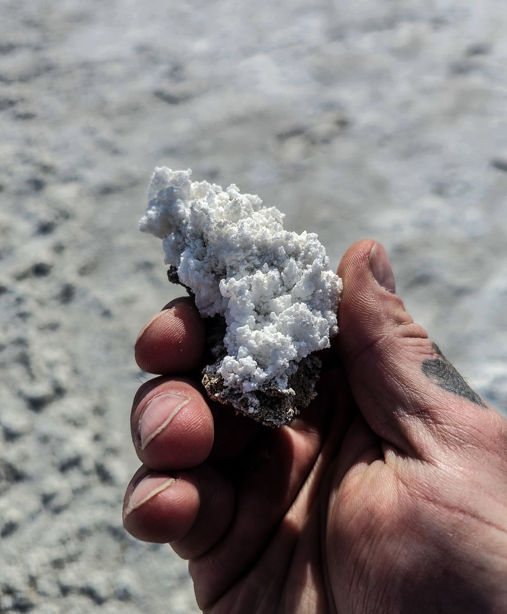 Hand holding a white crystalline mineral rock specimen against a blurred gray rocky background outdoors.