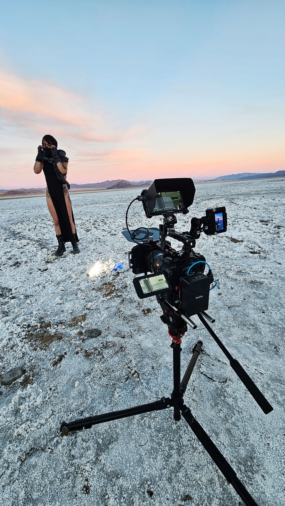 Professional camera rig on tripod filming a person in black outfit on a vast salt flat at sunset.