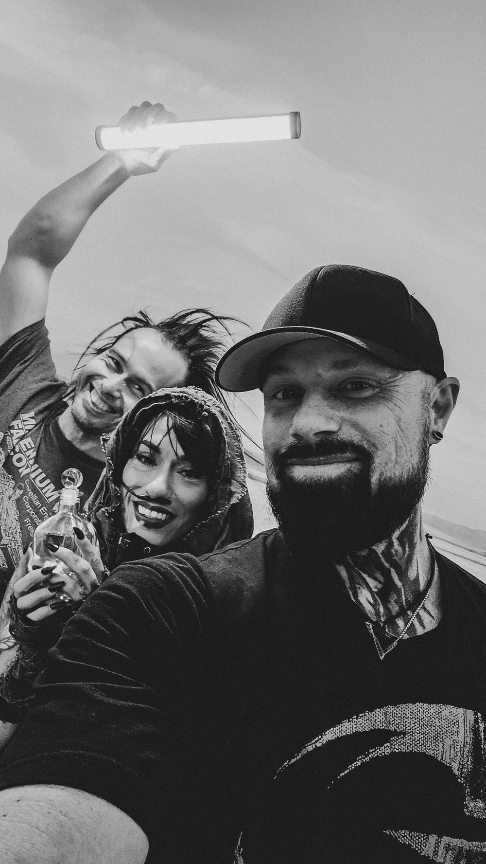 Black and white selfie of three smiling friends, one holding a glowing light stick, with tattoos and casual attire outdoors.