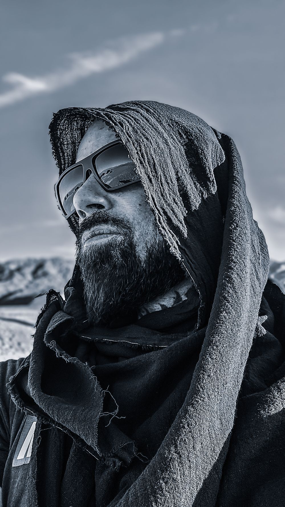 Black and white portrait of a bearded man wearing sunglasses and a wrapped scarf headcover in a snowy landscape.