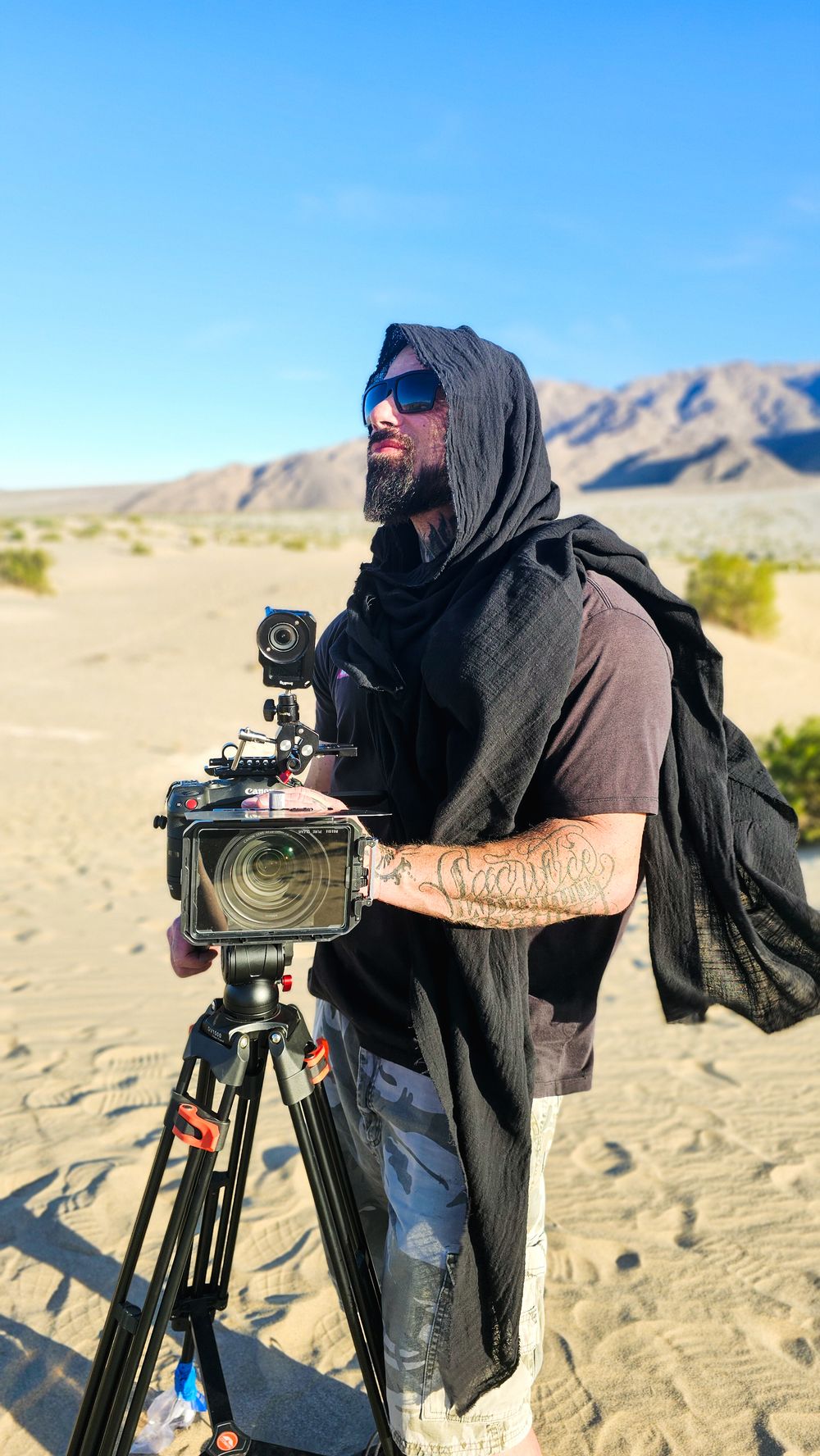 Bearded man with sunglasses and black hood stands beside a professional camera on tripod in a sandy desert landscape.
