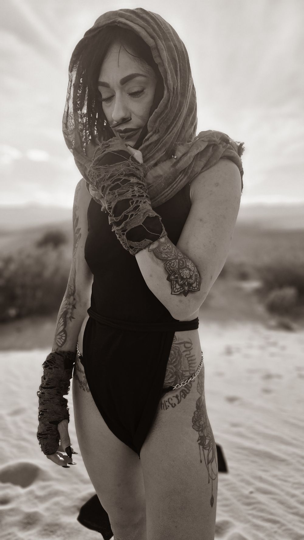 Black and white photo of tattooed woman in black swimsuit with lace head wrap posing in desert sand.
