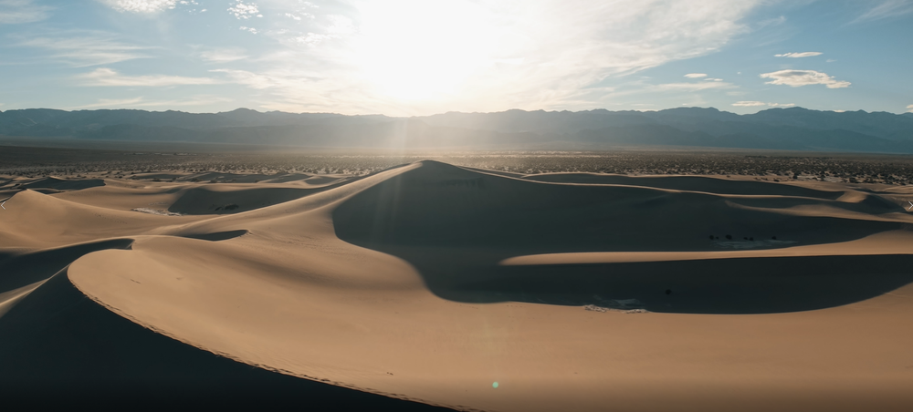 Aerial panoramic view of golden sand dunes in Death Valley desert with mountain range and bright sun on the horizon.