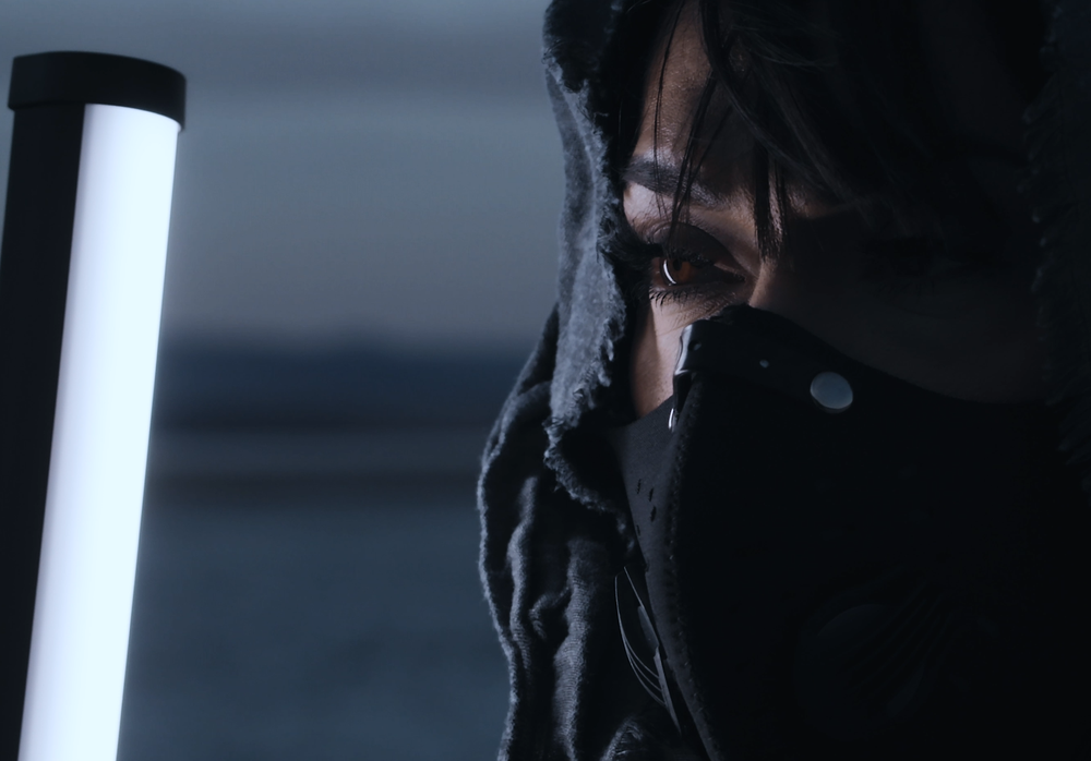 Person in dark hood holding a glowing white light tube in a dramatic, moody setting.