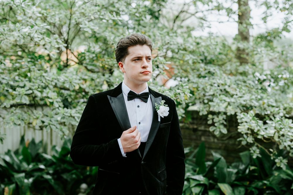 A person in a black tuxedo and bowtie stands in a lush garden setting holding a boutonniere.