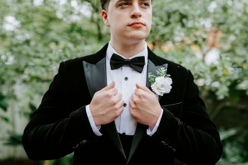 A person in a black tuxedo adjusts their bowtie while standing outdoors among green foliage.