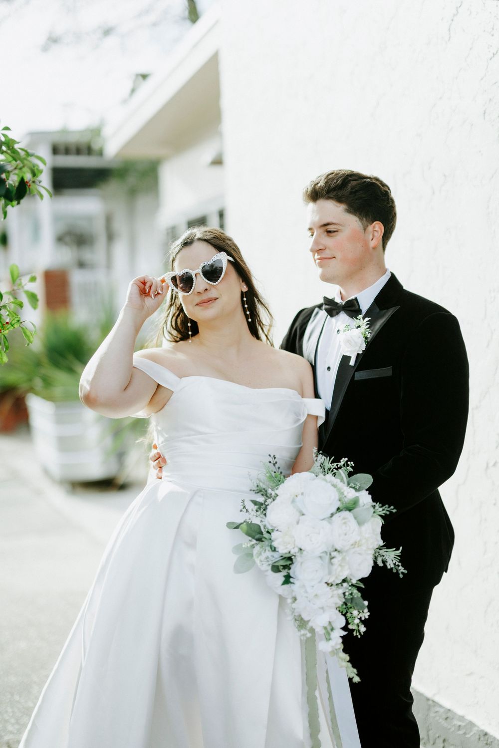 Elegant wedding couple in white dress and black tuxedo pose together in bright outdoor setting with floral bouquet.