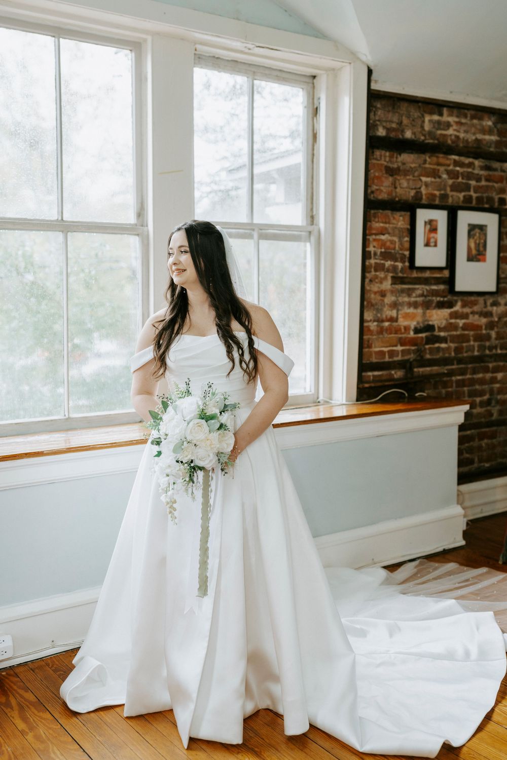 A bride in a flowing white gown twirls and poses in a room with exposed brick walls and large windows.