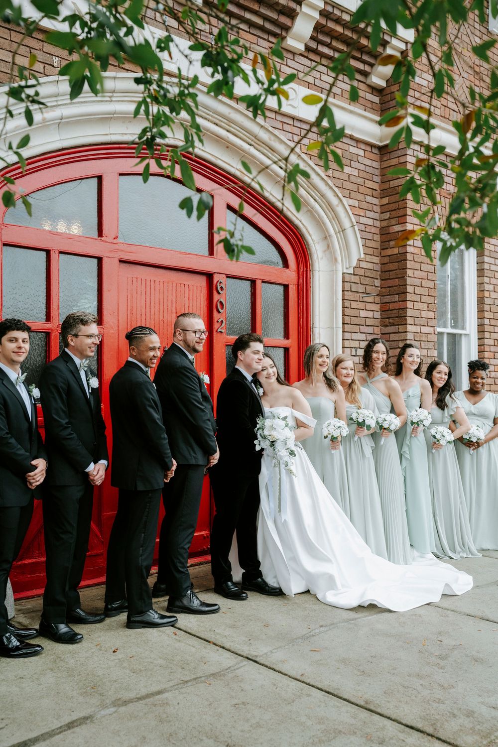 Wedding party lined up against red doors of brick building with bridesmaids in light blue and groomsmen in black suits.
