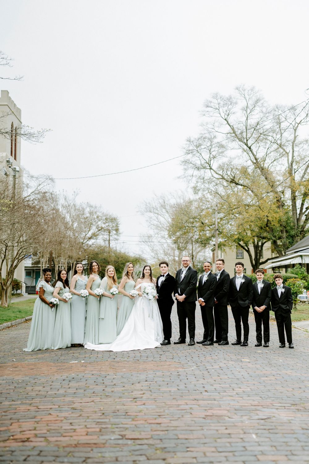 Wedding party in formal attire poses on cobblestone street with church spire visible in background.