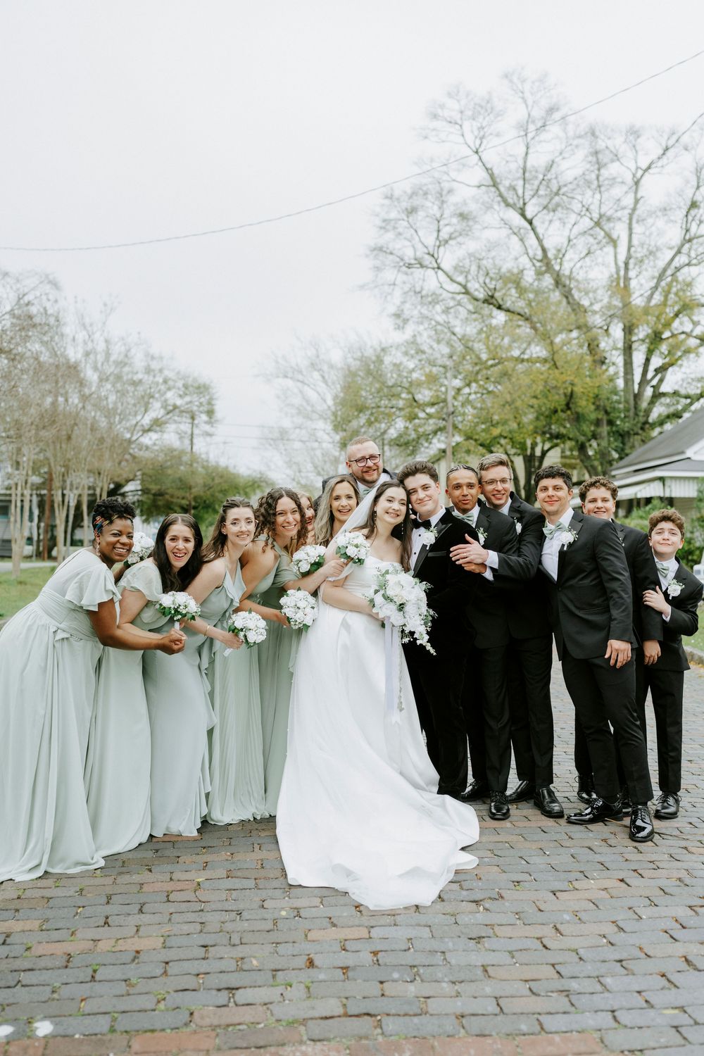Wedding party in light gray dresses and black suits poses together on brick pathway.