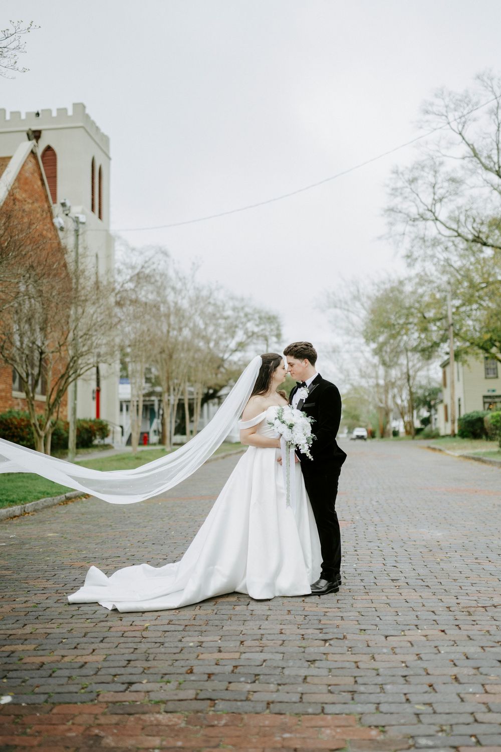 Bride and groom share intimate moments on historic cobblestone street with church steeple in background.